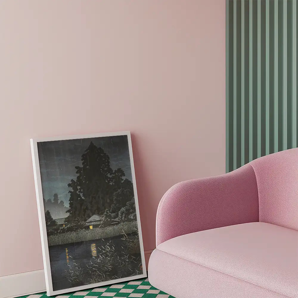 Framed painting of a serene landscape depicting a house by a river at night, placed against a pink wall next to a modern pink couch.