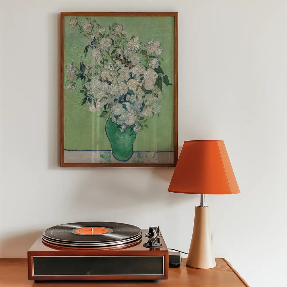 Painting of white flowers in a green vase against a light green background, framed and displayed above a vintage record player and an orange lampshade.