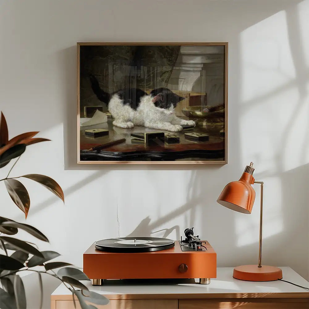 Framed painting of a black and white cat lying on a table surrounded by books, displayed above a retro turntable and a modern desk lamp in a sunlit room.