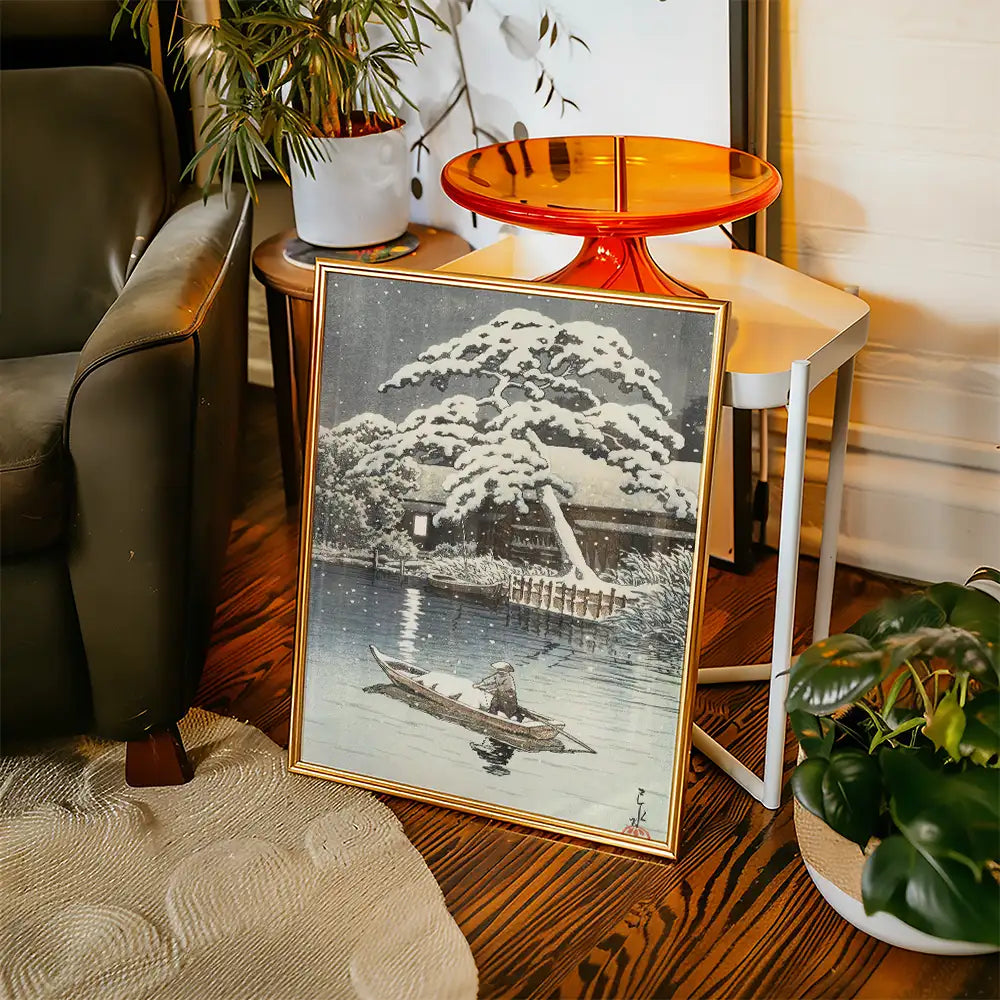 Framed painting of a serene winter scene depicting a person in a boat on a river with snow-covered trees and a house in the background, placed next to a modern side table and plants.