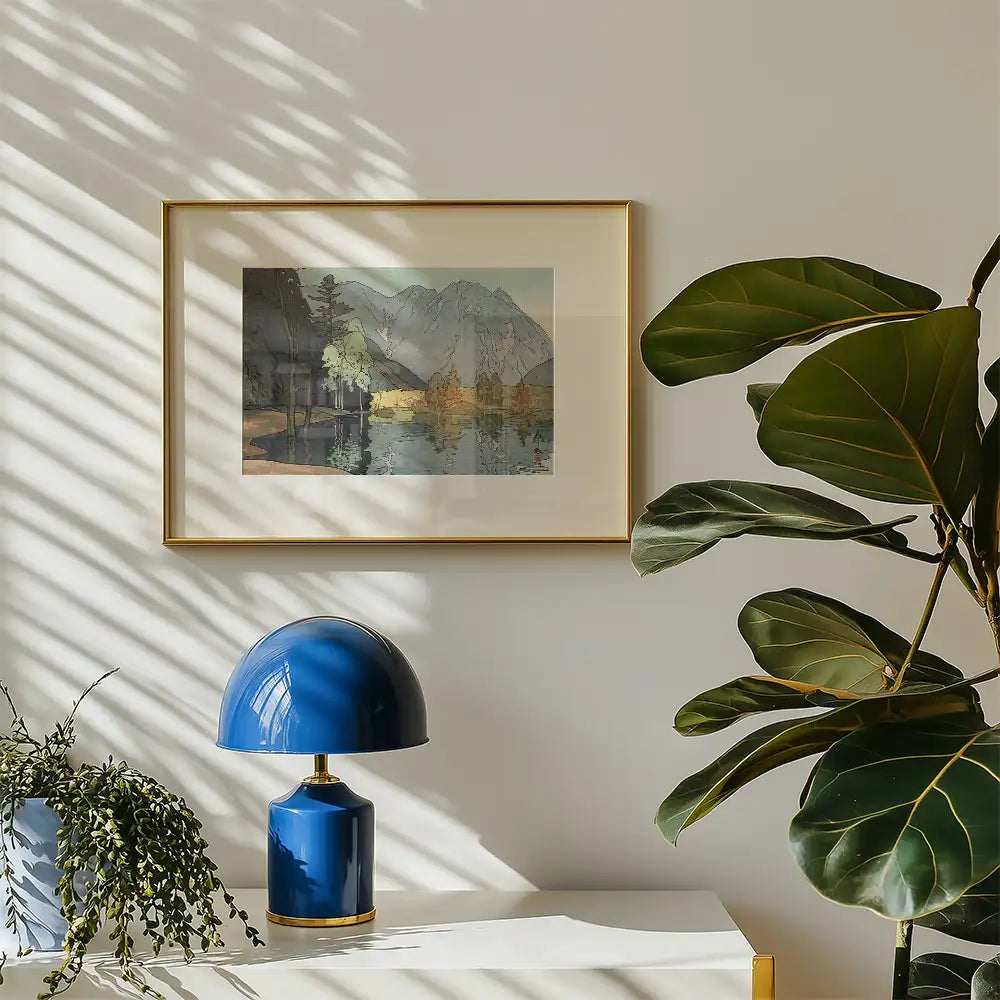 Framed painting of a serene landscape with mountains and trees, displayed on a light wall beside a blue lamp and a leafy plant.