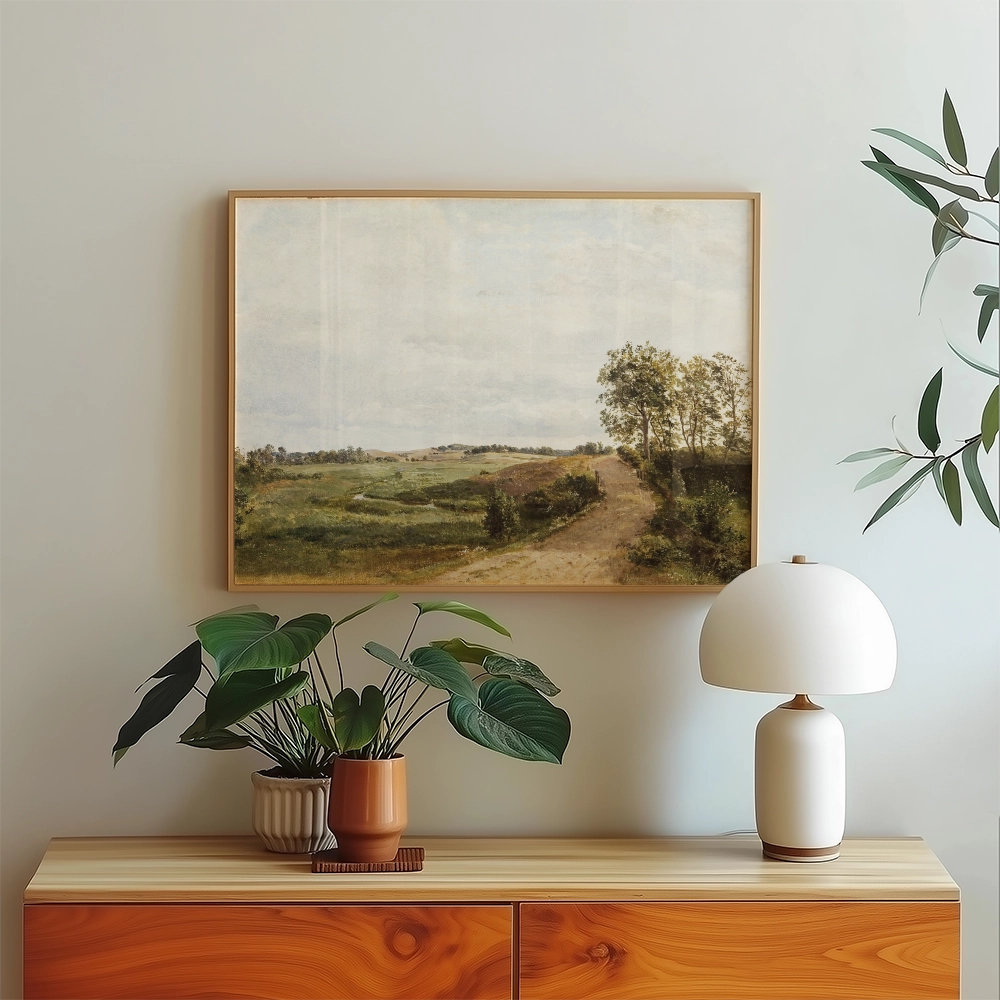 Framed painting of a scenic landscape featuring green fields, a winding path, and trees, displayed above a wooden sideboard with a plant and lamp.