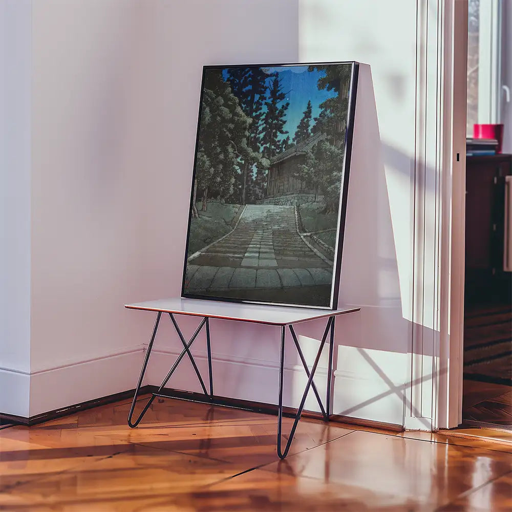 A framed painting of a stone pathway leading to a wooden building, surrounded by green trees, displayed on a modern table in a well-lit room.