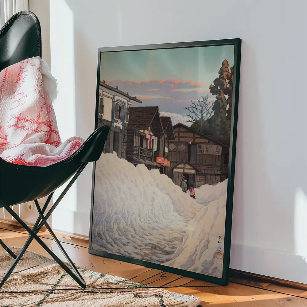 Framed painting depicting snow-covered houses and a figure in a winter landscape, leaning against a wall beside a black chair and a cozy blanket.