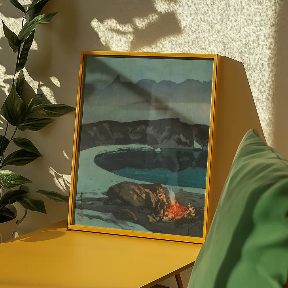 Framed painting of a serene landscape featuring a lake and mountains, displayed on a yellow table with a green pillow and plants in the background.