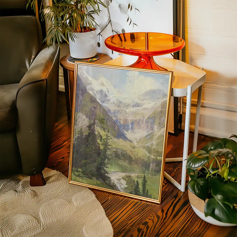Framed painting of a mountainous landscape displayed in a cozy interior setting with a green plant and a small orange table.