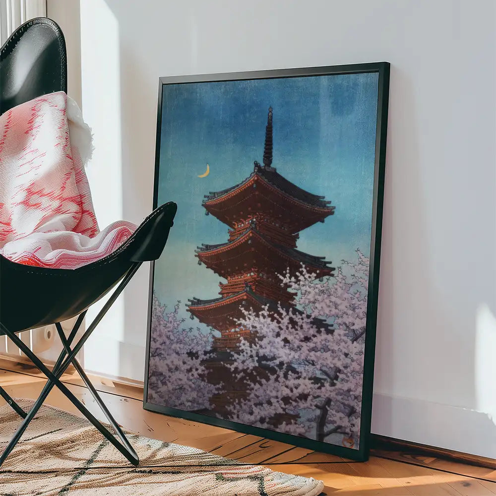 Framed painting of a traditional pagoda surrounded by cherry blossoms, leaning against a wall beside a black chair with a blanket draped over it.