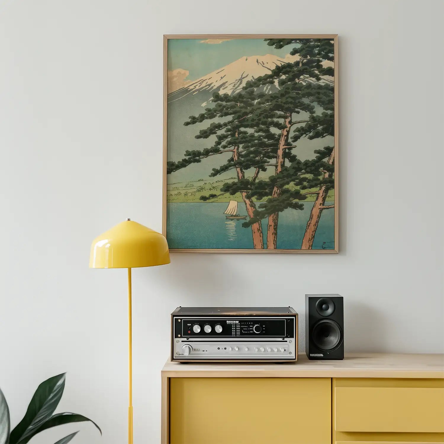 Framed painting of a mountain landscape featuring trees, a lake, and a sailboat, displayed above a yellow lamp and vintage stereo speaker on a modern sideboard.