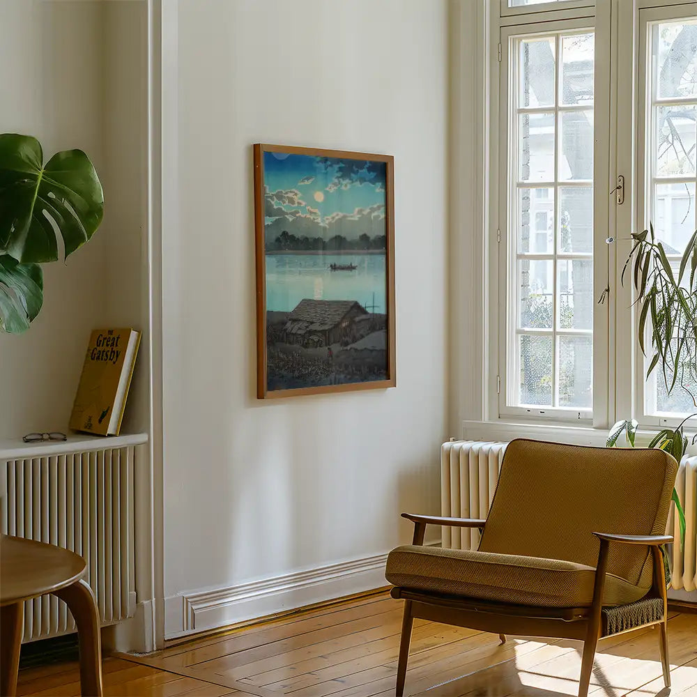 Framed painting of a serene riverside scene depicting a boat and a house, hanging on a white wall in a cozy living room with a chair and a plant.