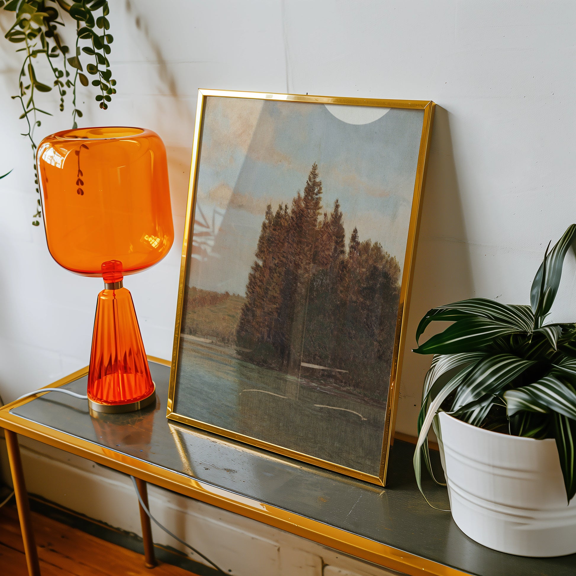 Framed landscape painting of trees beside a calm body of water, displayed on a table alongside an orange lamp and a potted plant.