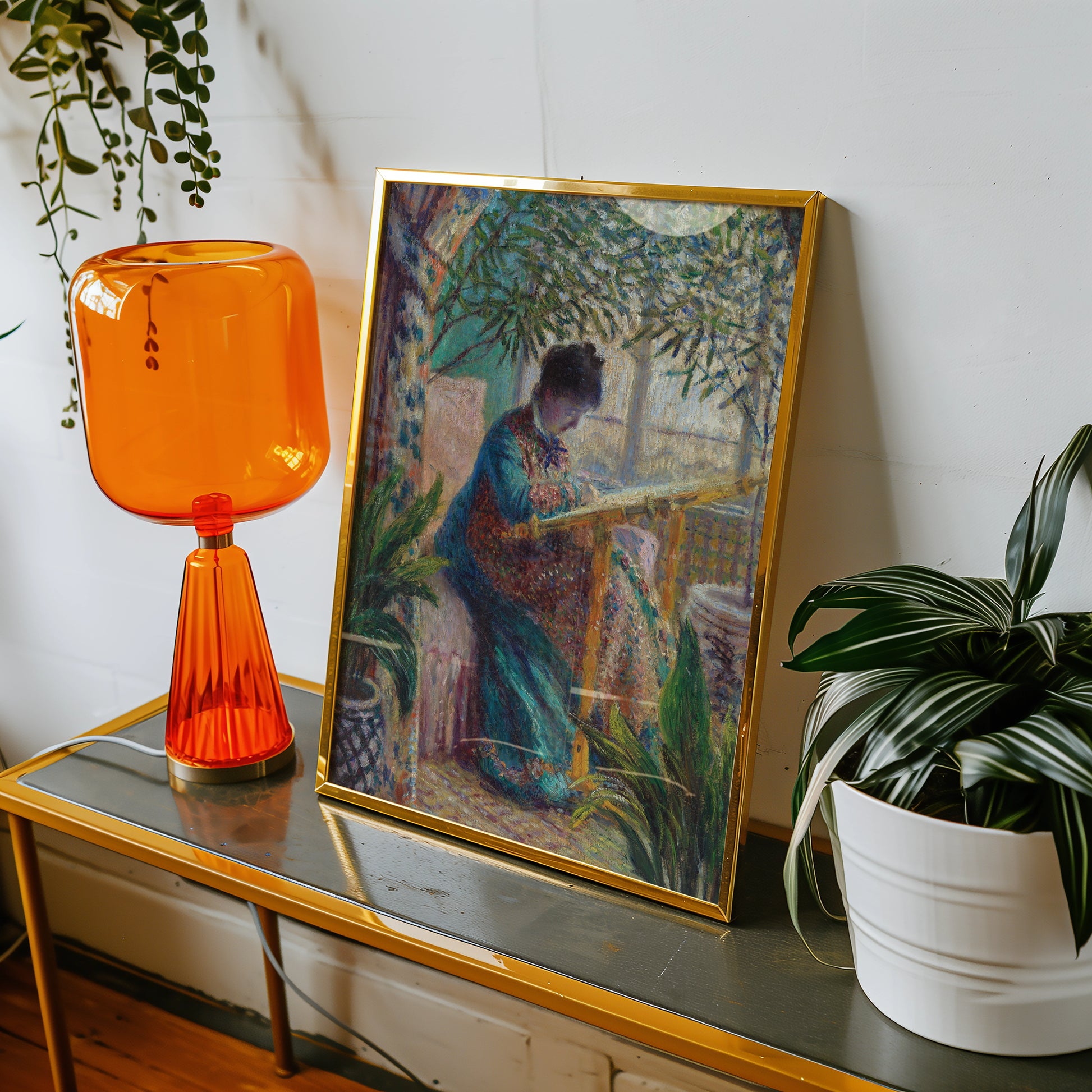 Painting of a woman seated in a colorful outfit, engrossed in an artistic activity, displayed in a gold frame next to an orange lamp and potted plant.