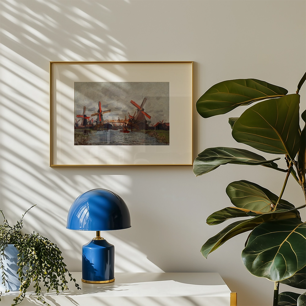 Framed painting of windmills in a landscape setting on a wall, accompanied by a blue lamp and a leafy plant on a white table.