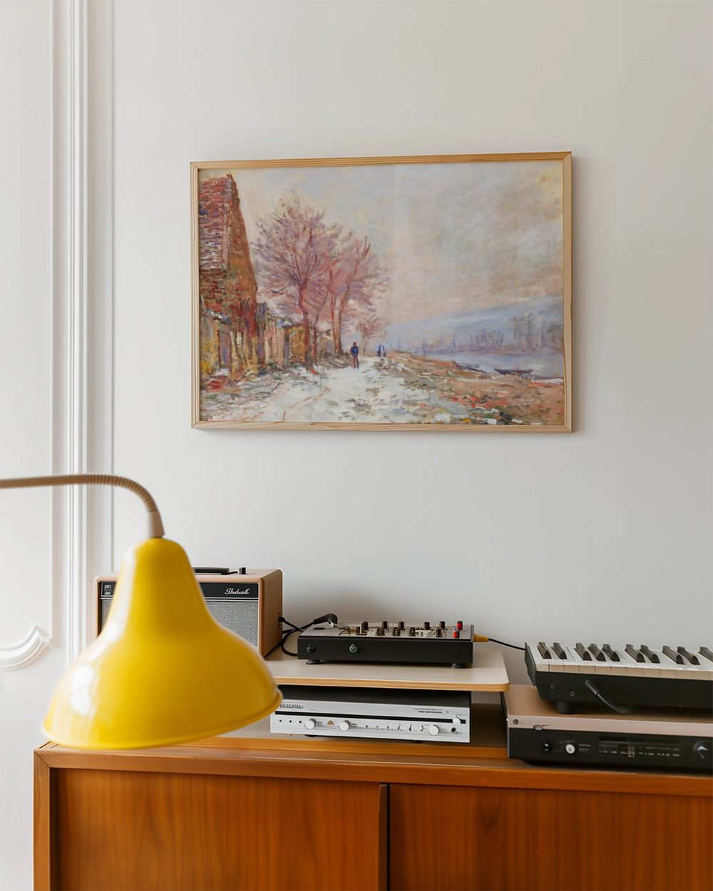 Framed painting of a serene landscape with trees and two figures walking along a path, hanging on a light wall above a wooden sideboard with a yellow lamp and audio equipment.
