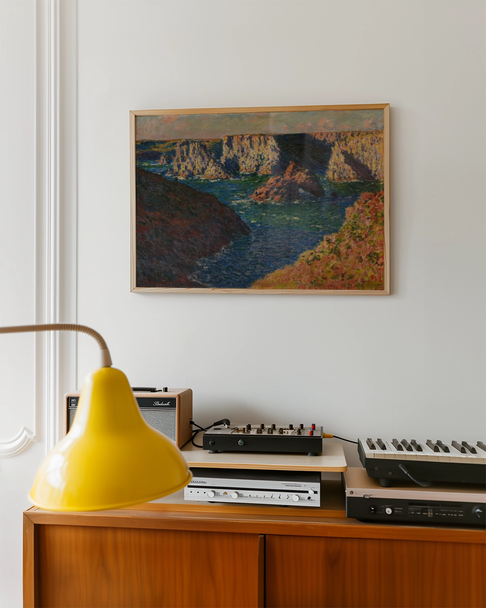 Painting of a serene landscape with cliffs and water, framed and displayed on a white wall above a vintage audio setup and a yellow lamp.