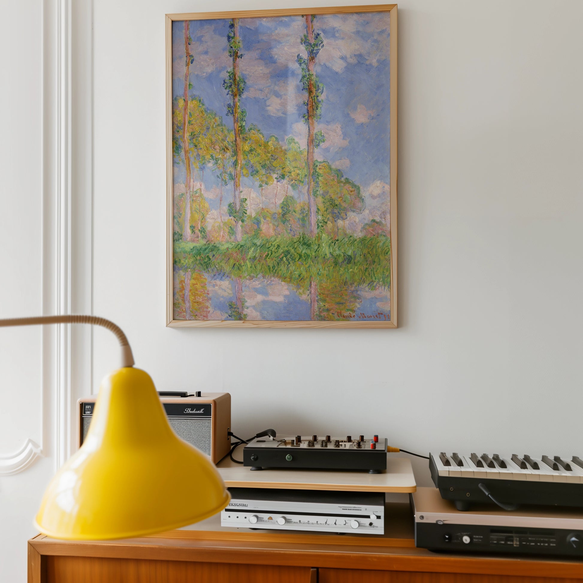 Painting of tall trees and blue sky with reflections in water, framed and displayed on a wall above a side table featuring a yellow lamp and various audio equipment.