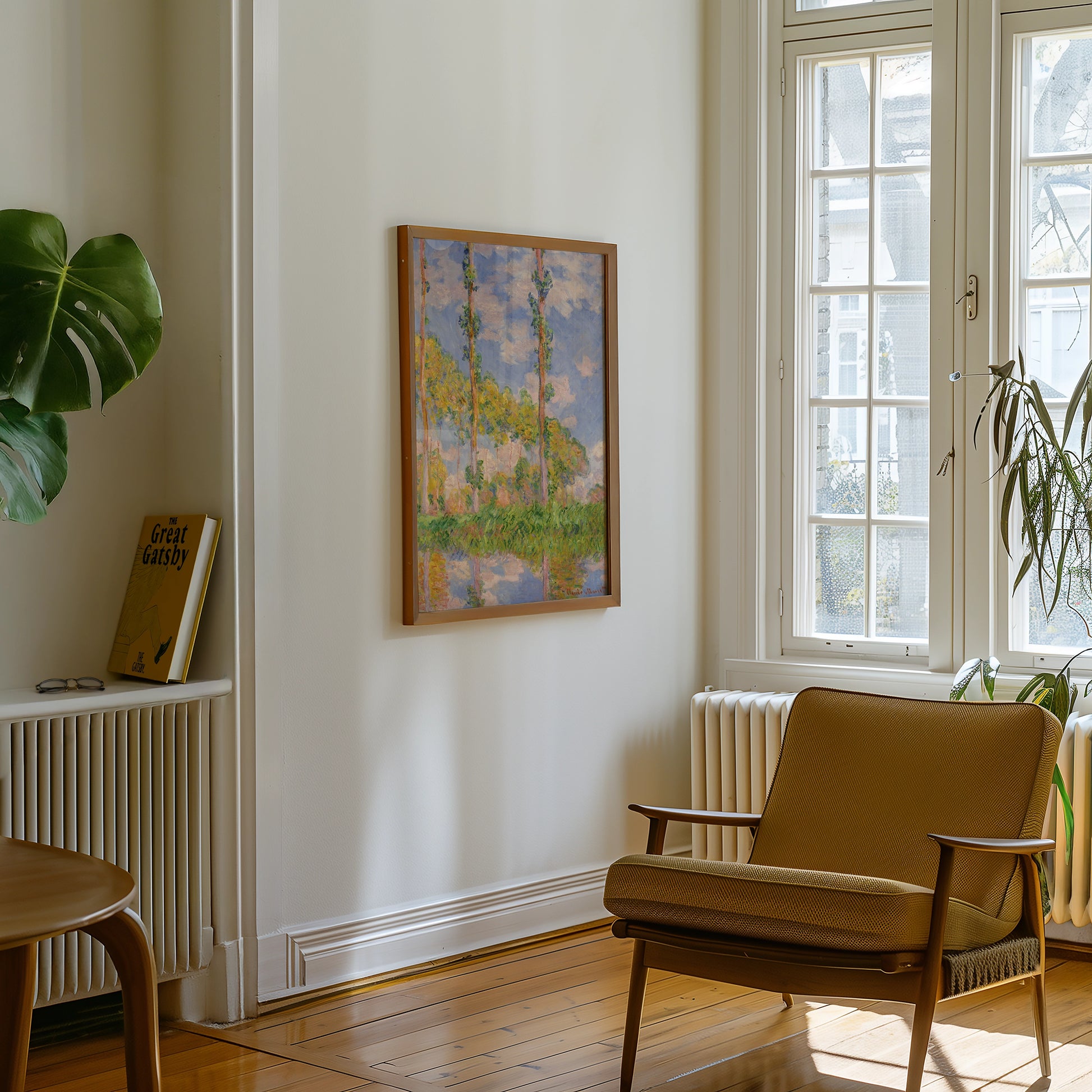 Painting framed and hanging on a white wall in a cozy room with a brown armchair, plant, and a book on a shelf.