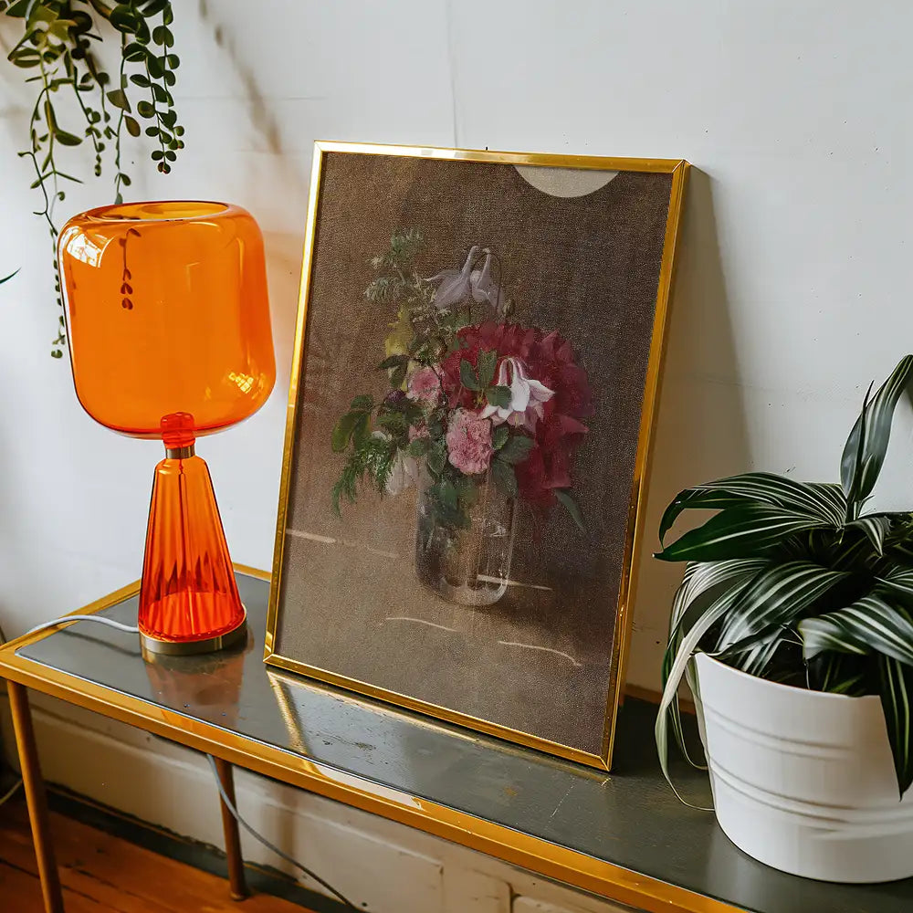 A framed painting of a floral arrangement featuring pink and white flowers, displayed on a gold-colored table beside an orange lamp and a potted plant.