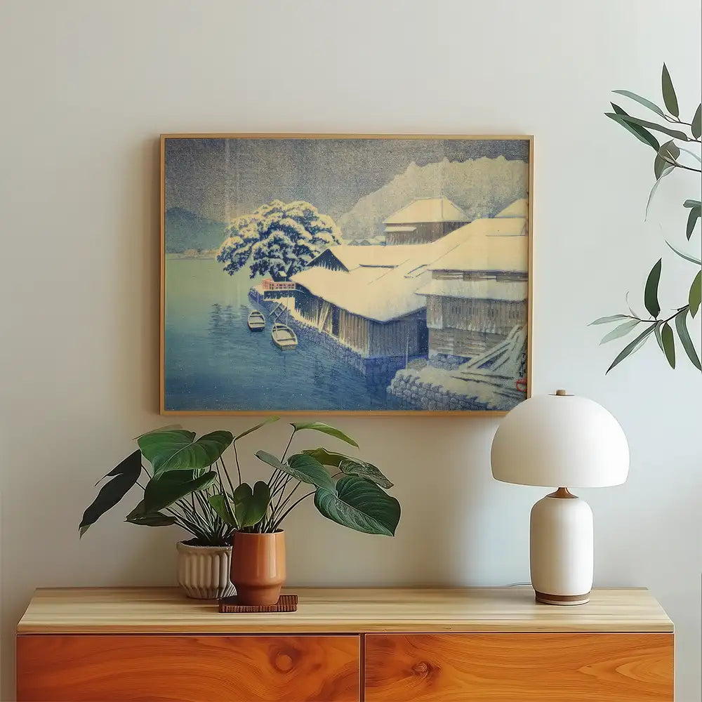 Framed painting of a winter landscape with snow-covered houses and boats by a calm waterway, displayed above a wooden sideboard with a potted plant and a lamp.