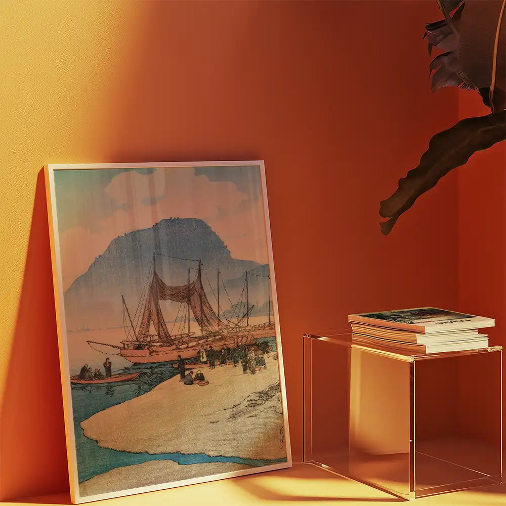 Framed painting of a coastal scene with boats and a mountain backdrop, displayed against a vibrant orange wall, accompanied by stacked books on a clear acrylic table.