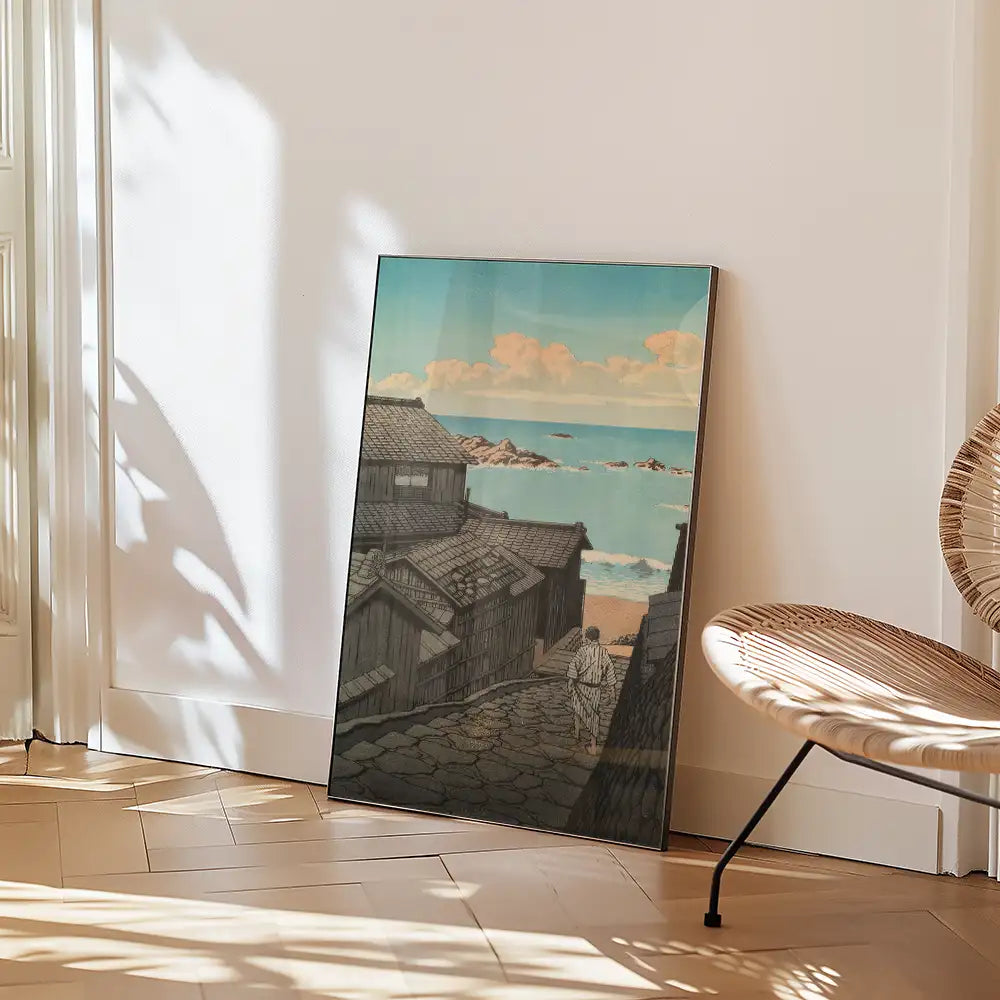 Framed painting of a coastal village scene with traditional houses and a beach, leaning against a wall in a bright room with a modern chair nearby.