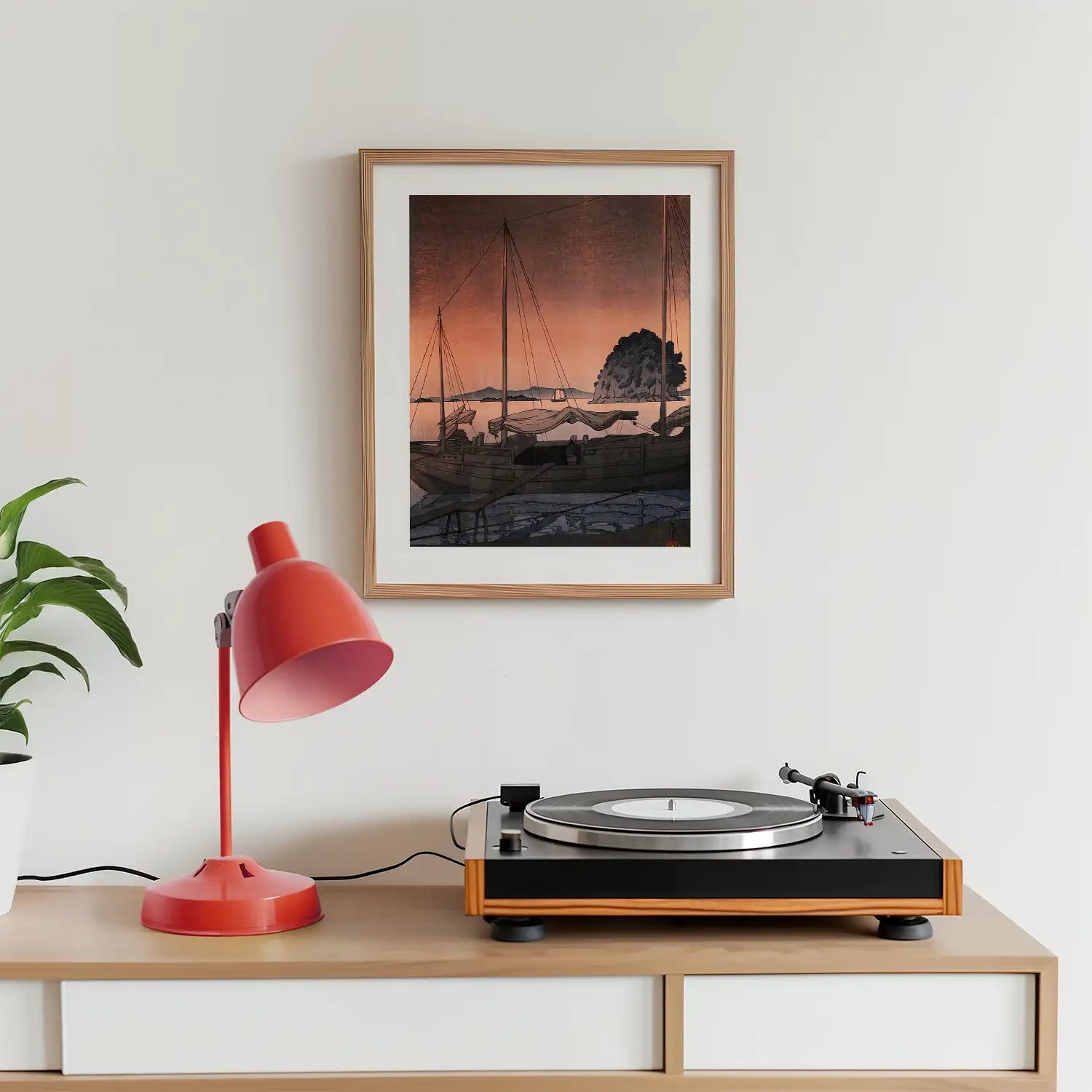 Framed painting of sailboats against a sunset, displayed above a record player and a red desk lamp on a wooden dresser.