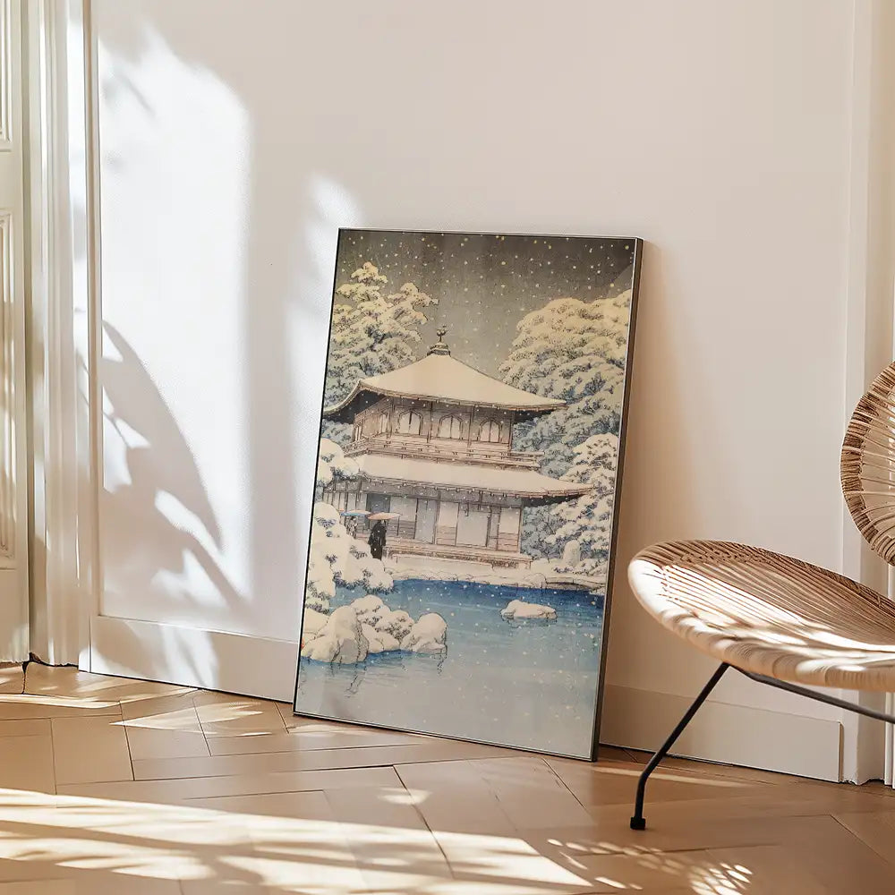 Painting of a snow-covered traditional Japanese house beside a tranquil pond, leaning against a wall in a bright, minimalistic interior setting.