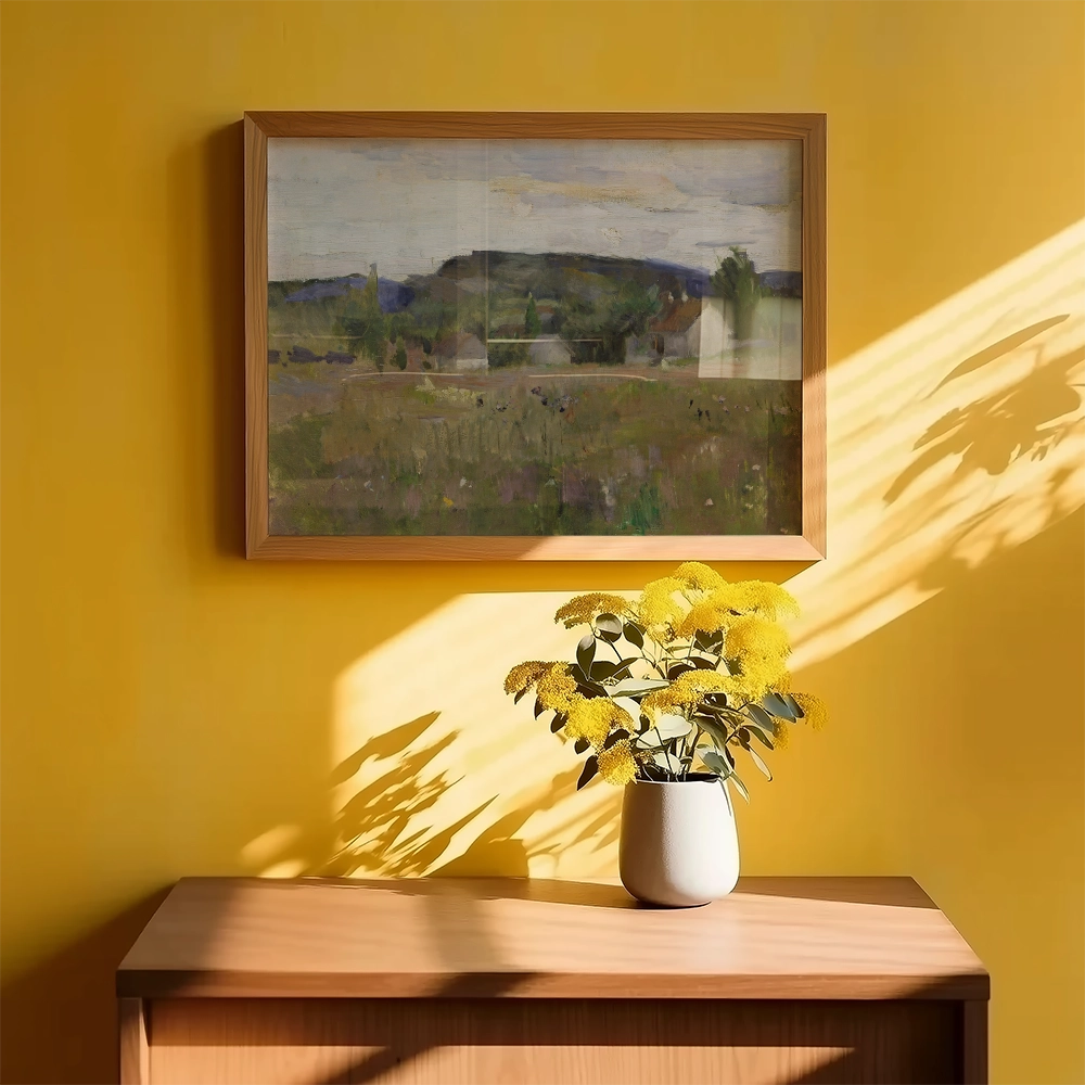 Framed landscape painting hanging on a yellow wall above a wooden table with a vase of yellow flowers.