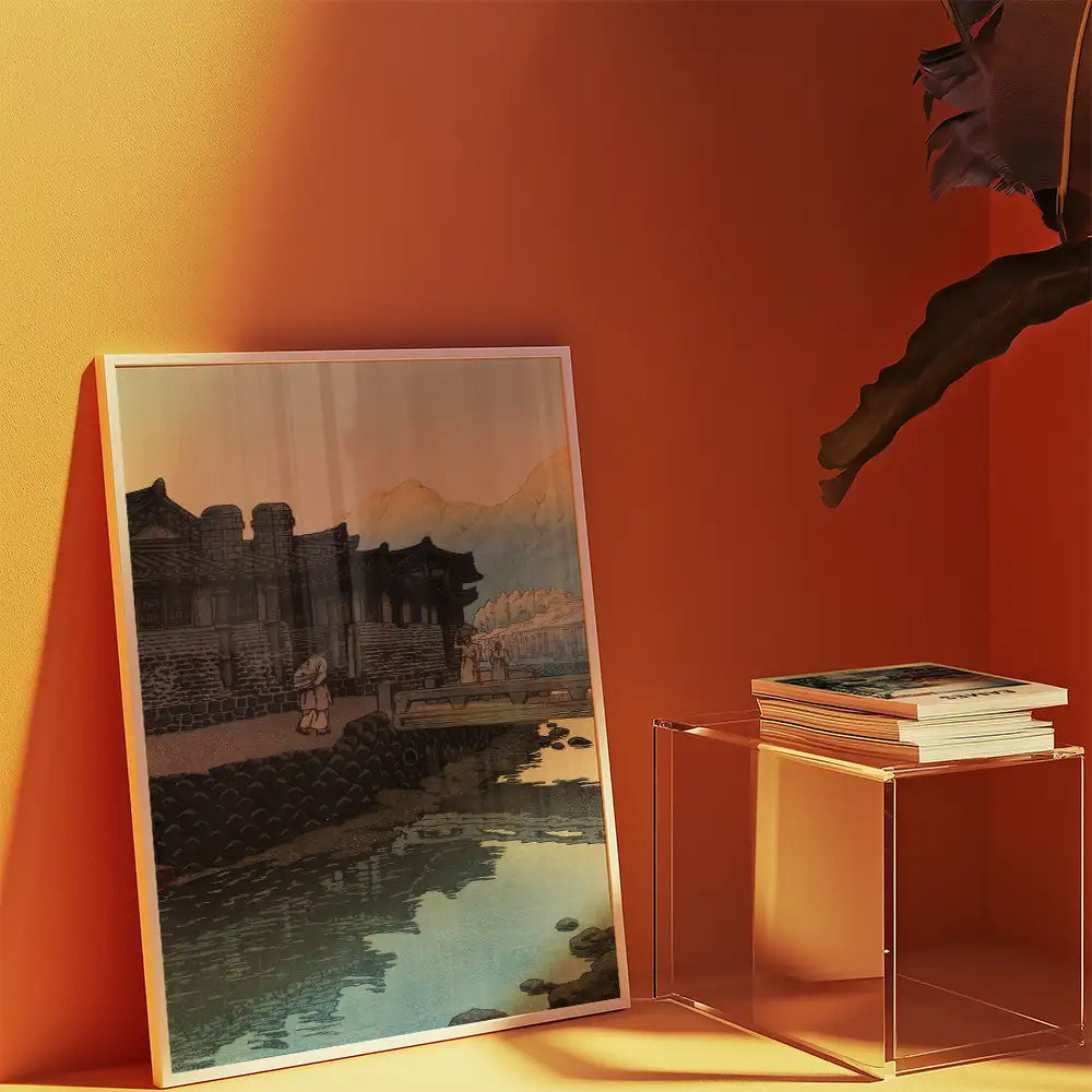 Framed painting of a serene landscape with a river, mountains, and traditional buildings, displayed against an orange wall with a clear side table and stacked books.