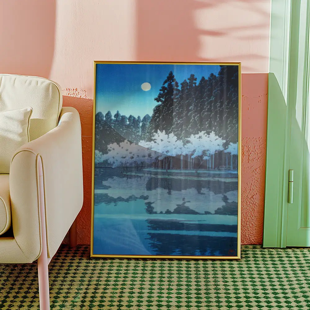 Framed painting of a serene landscape featuring a moonlit lake and forest in shades of blue, displayed next to a light-colored armchair in a stylish interior setting.