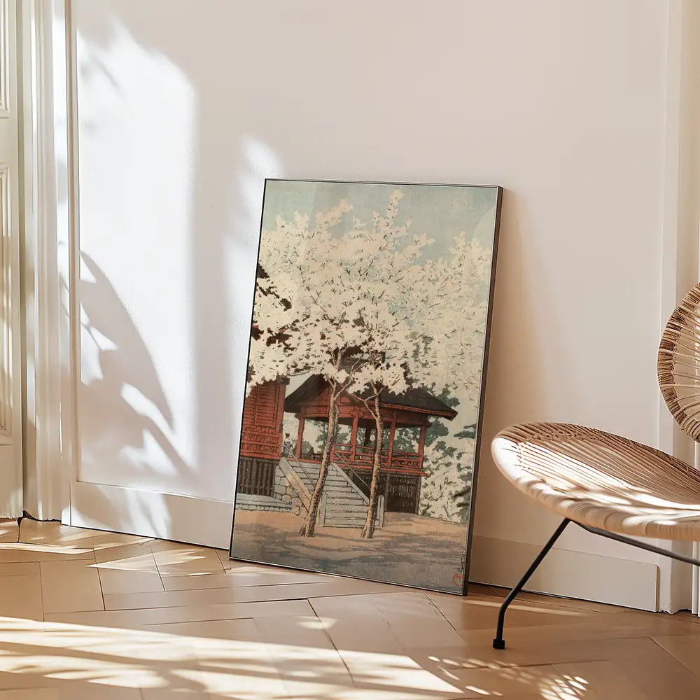 Framed painting of a traditional building surrounded by blooming trees, leaning against a white wall in a stylish interior setting with a woven chair nearby.