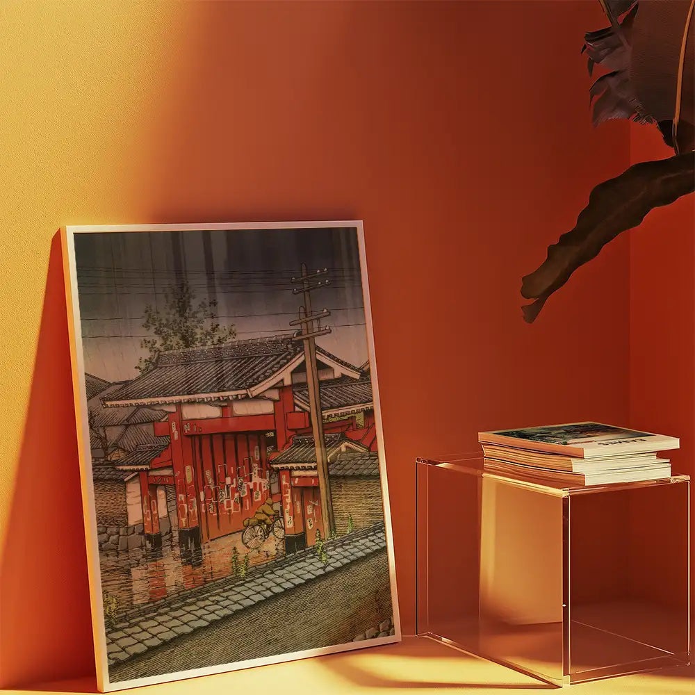 A colorful painting of a traditional Japanese street scene featuring a red building and a bicycle, displayed against an orange wall with a clear, modern table holding stacked books.