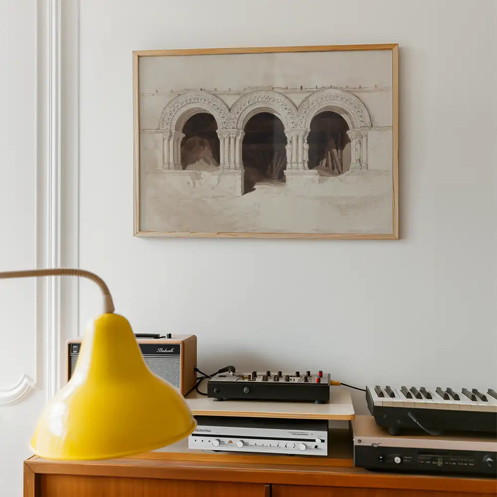 Framed painting of arched architectural features hanging on a white wall, with a yellow lamp and audio equipment in the foreground.