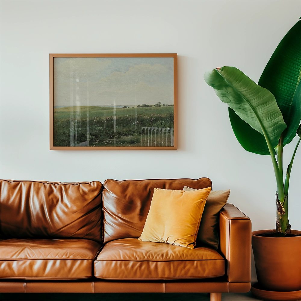 A framed landscape painting depicting a serene green field, hanging above a stylish brown leather couch with decorative pillows and a large green plant beside it.