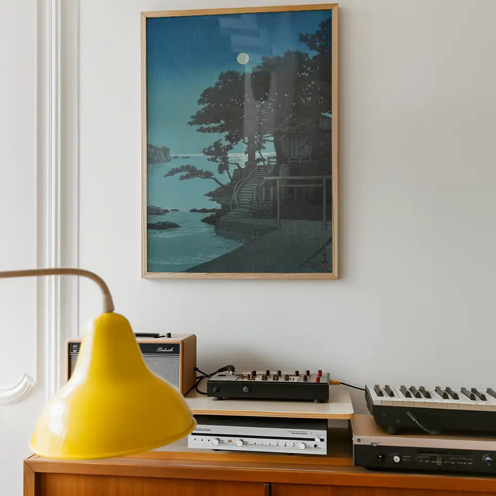 Framed painting of a moonlit landscape featuring trees and a staircase by a river, displayed above a wooden table with a yellow lamp and audio equipment.