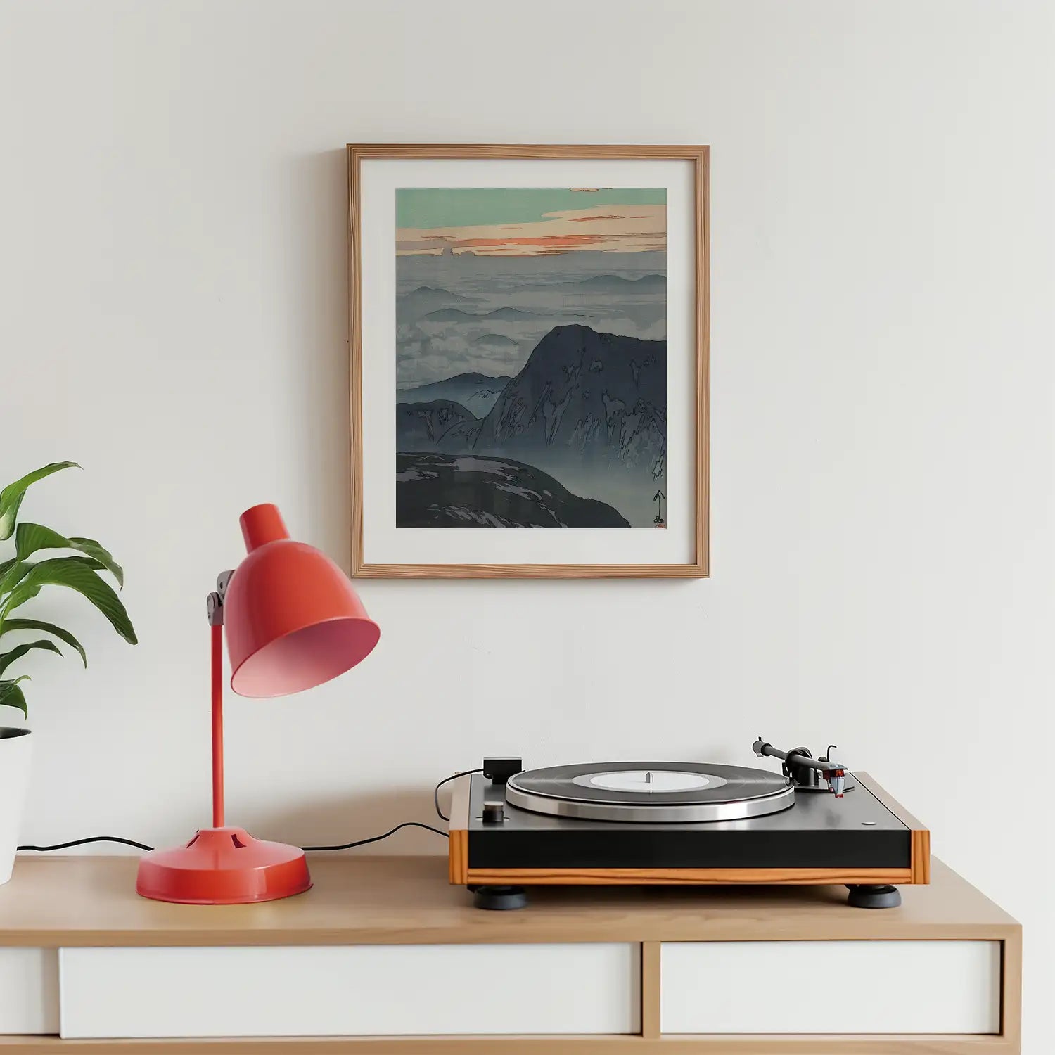 Framed mountain landscape painting on a wall above a record player and a red desk lamp in a modern living space.