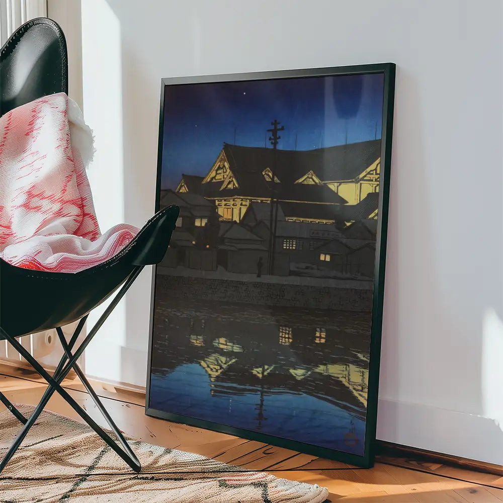 Framed painting of a nighttime scene depicting illuminated buildings reflecting in water, placed next to a black chair with a cozy blanket.