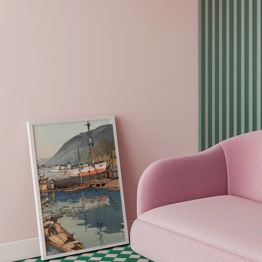 A framed painting of a harbor scene featuring boats and mountains, leaning against a pastel pink wall next to a pink sofa.