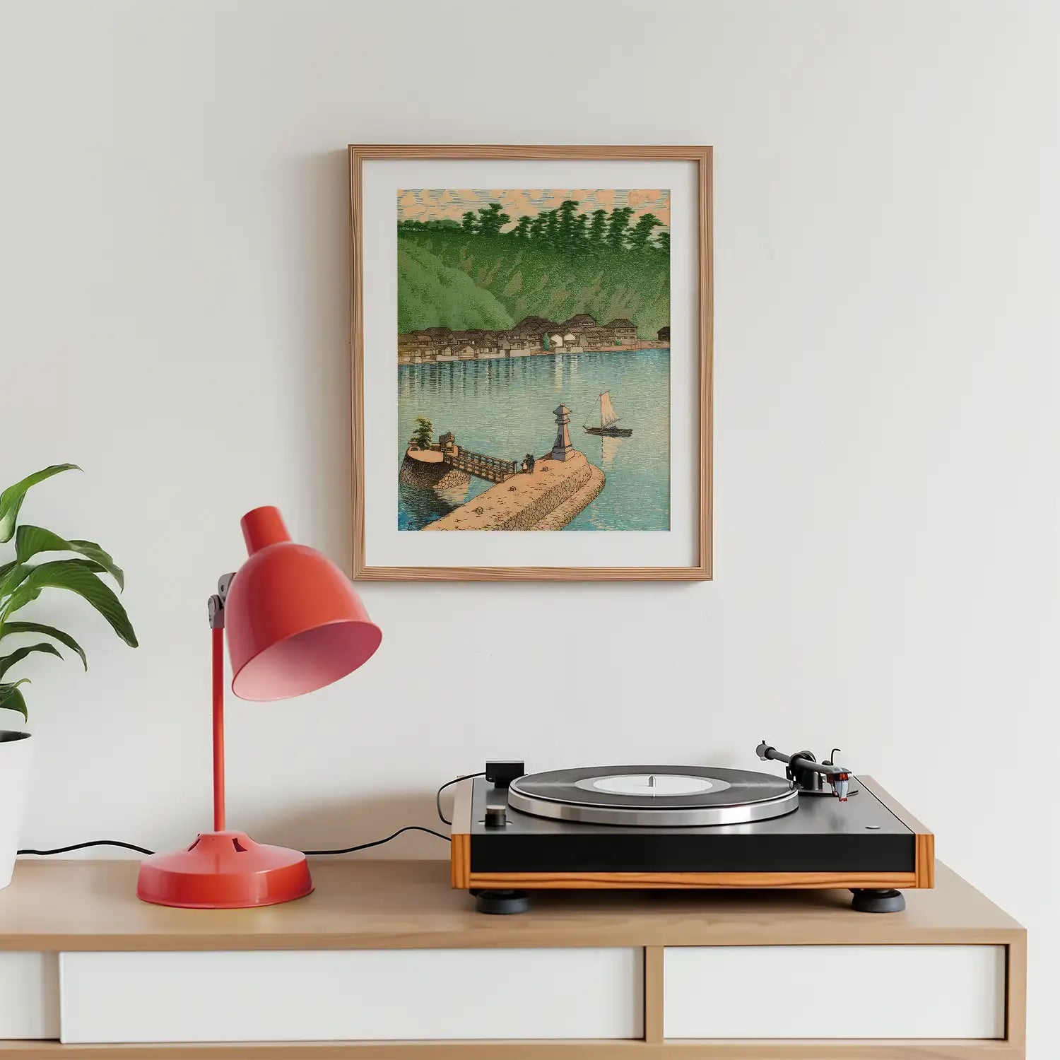 Framed painting of a coastal scene with a lighthouse and sailboat, displayed above a wooden table featuring a record player and a red desk lamp.