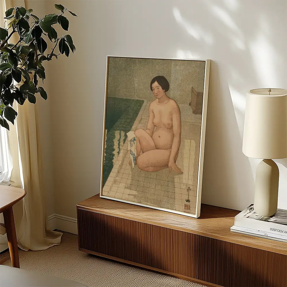 A framed painting of a nude woman sitting by a pool, displayed on a wooden console table in a well-lit room with a lamp and plant.