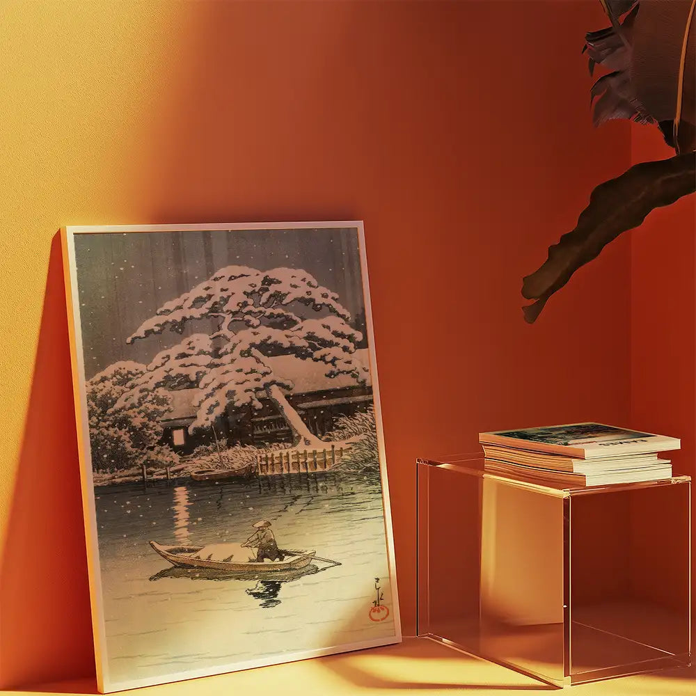 Painting of a tranquil winter scene featuring a snow-covered tree and a person in a boat on a calm lake, displayed against a warm orange wall with a clear acrylic side table and stacked books.
