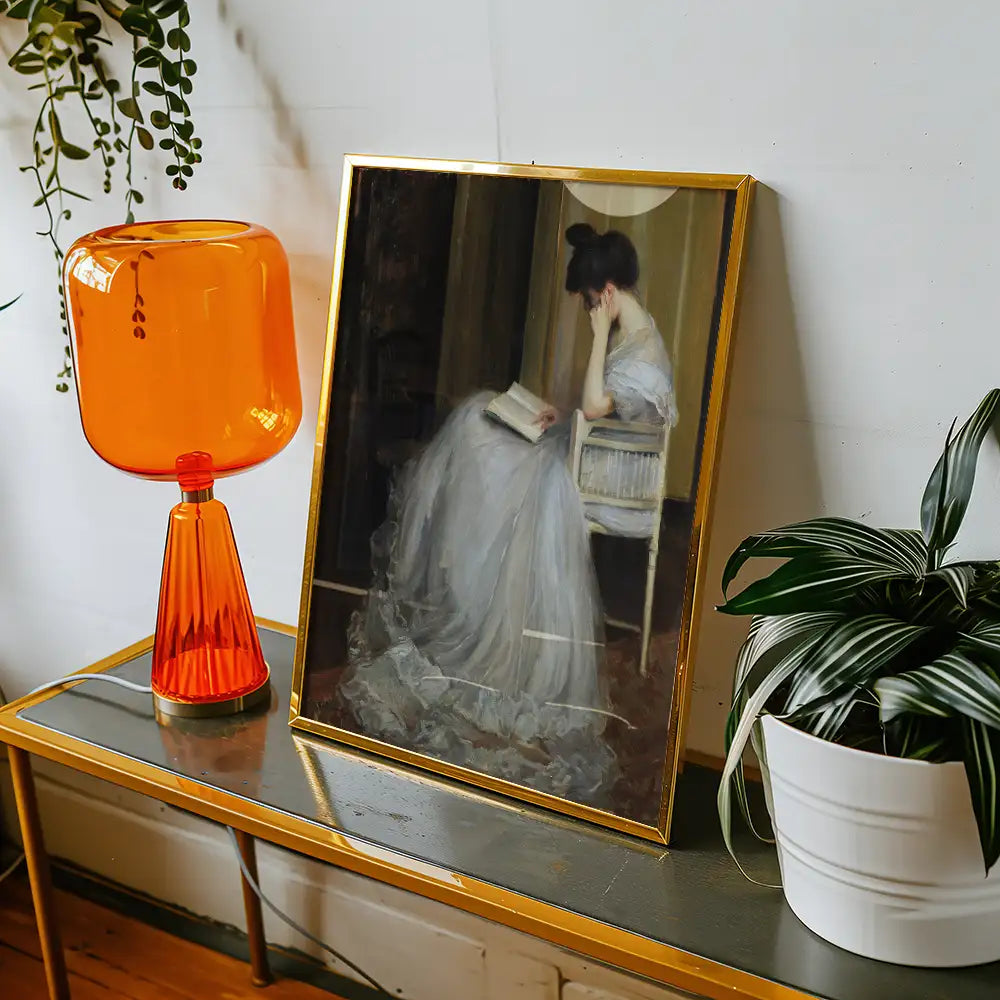 Portrait of a woman in a flowing gray dress reading a book, framed in gold, displayed beside an orange lamp and a plant on a table.