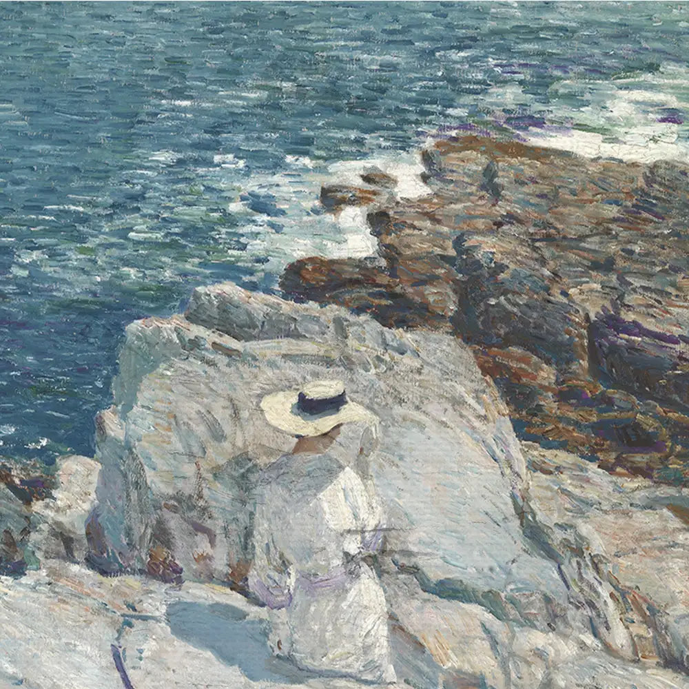 A woman in a white dress and straw hat sitting on a rocky shore, gazing out at the ocean.