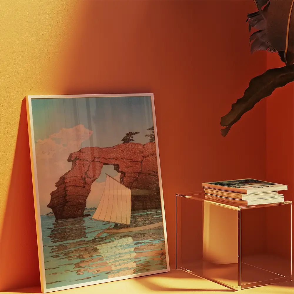 Framed painting of a sailboat near a rock arch, displayed in a warm-toned interior with a clear side table and stacked art books.