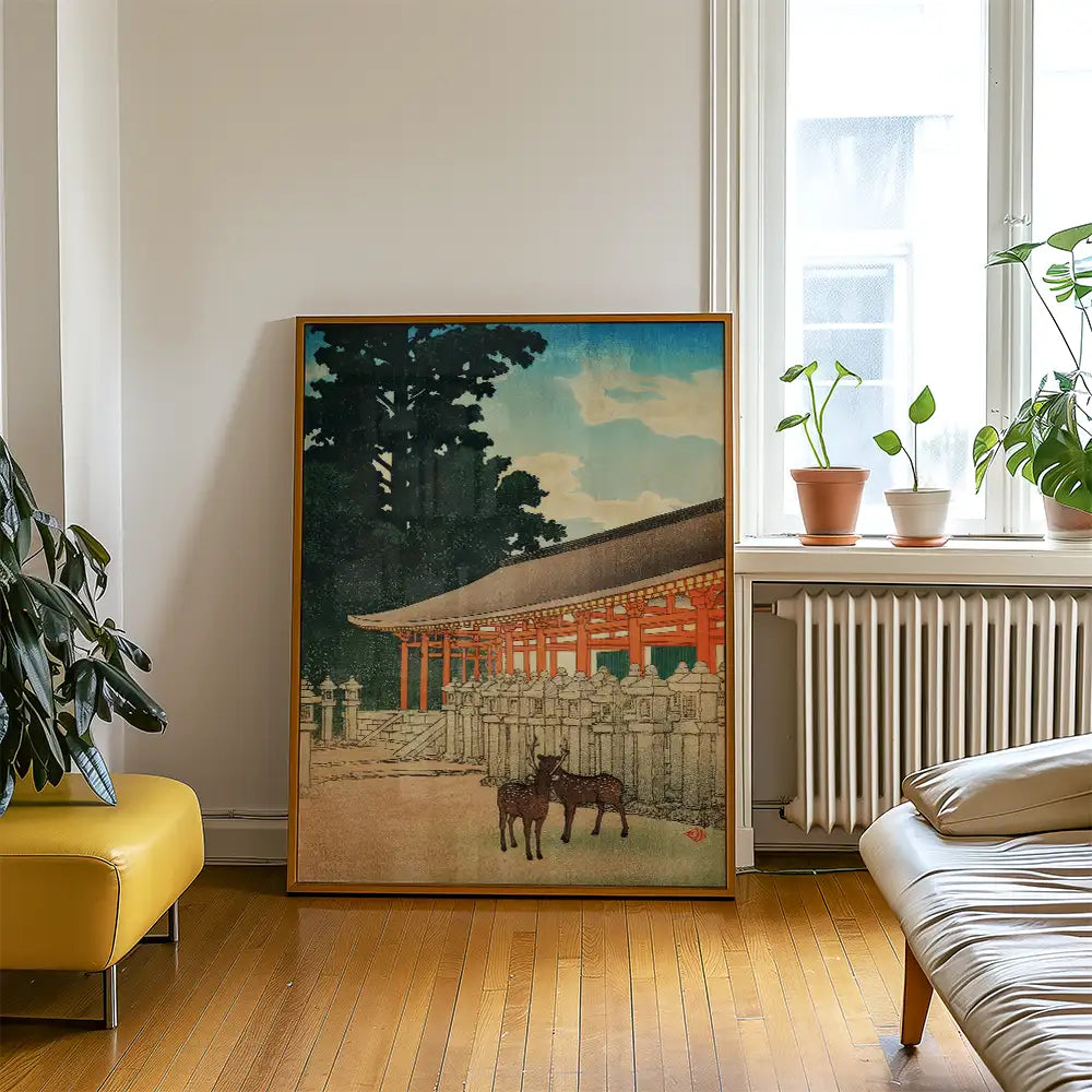 Painting of a serene landscape featuring a traditional building and two deer, framed and displayed in a room with plants and natural light.