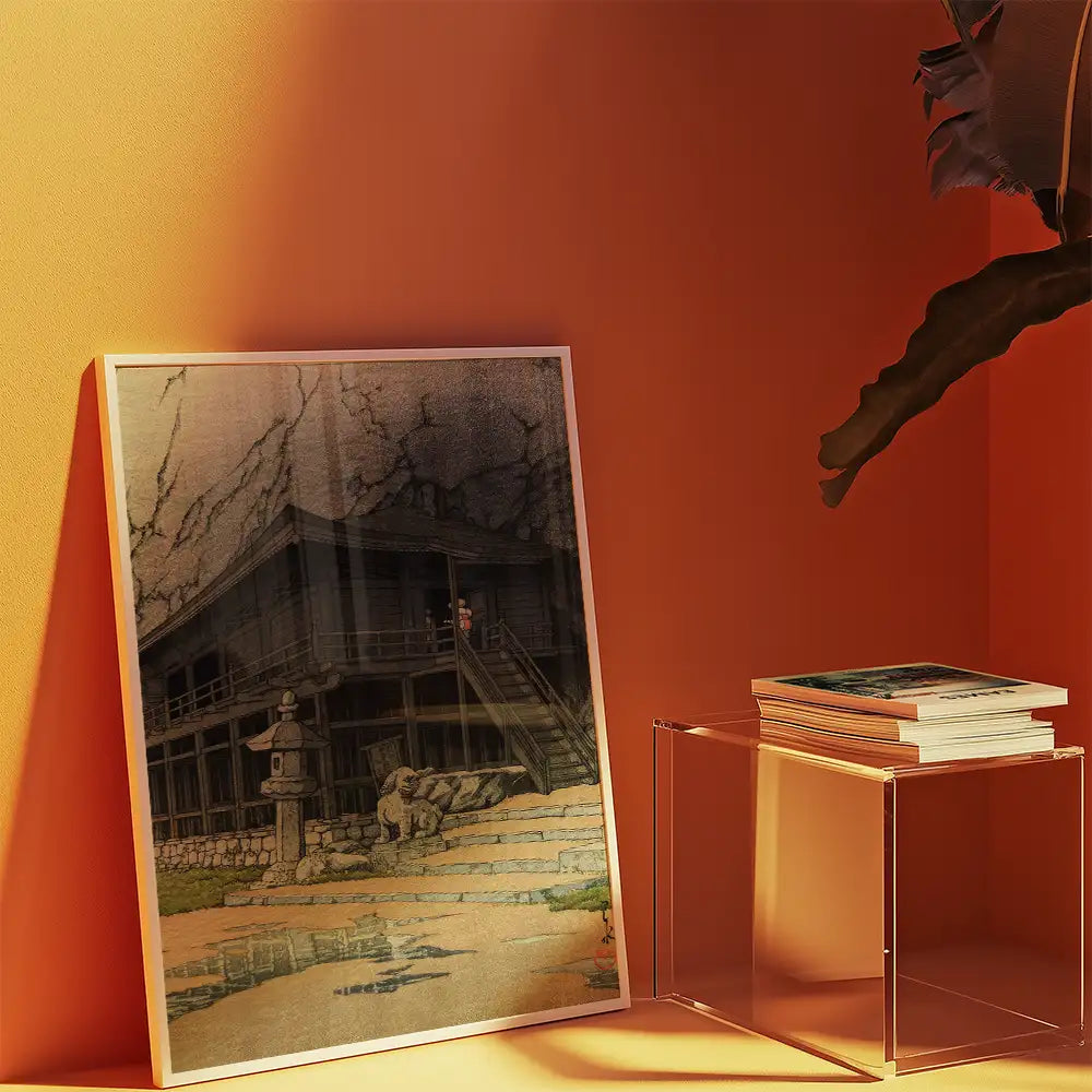 Framed painting of a traditional Japanese house displayed against an orange wall, with a stack of books on a clear side table.