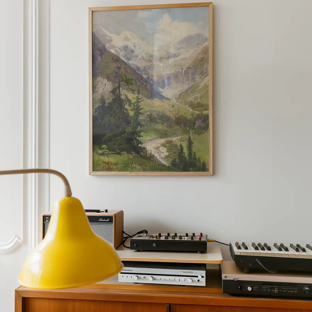 Framed painting of a mountainous landscape with green meadows and pine trees, displayed on a white wall above a wooden side table with a yellow lamp and sound equipment.