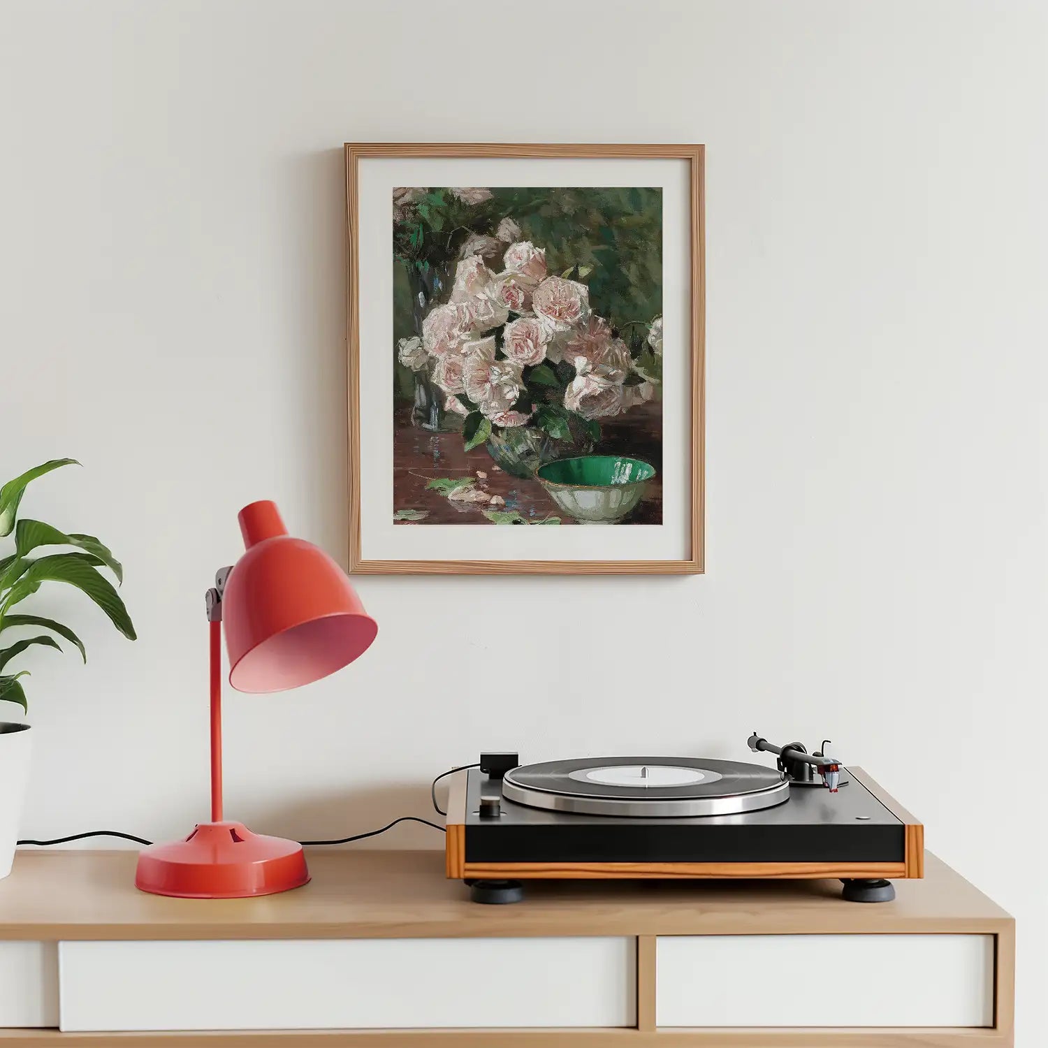 Framed painting of pink roses in a vase hanging above a modern record player and a red desk lamp on a wooden surface with a green bowl beside the vase.