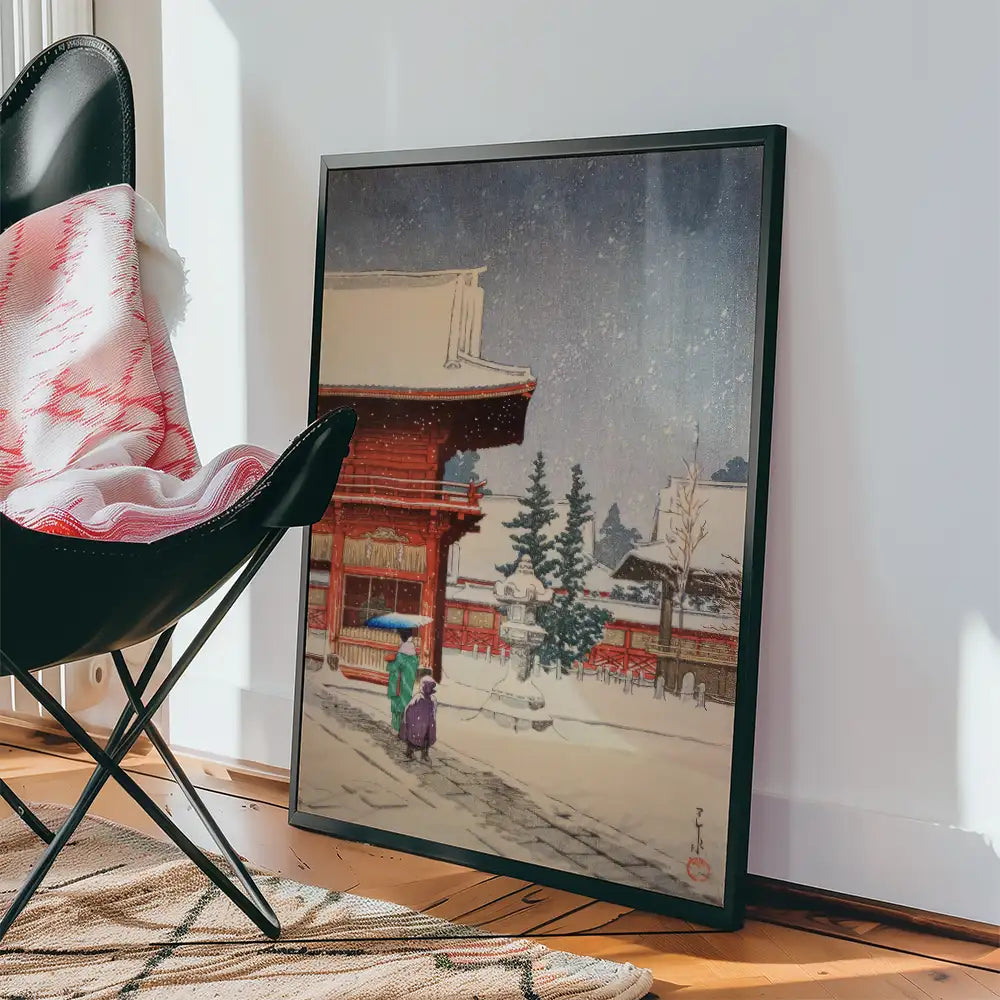 Framed painting of a snow-covered traditional Japanese temple scene with two figures walking under an umbrella, placed beside a black chair and a patterned rug.