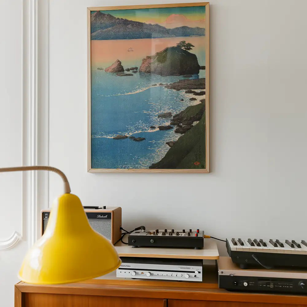 Framed painting of a coastal landscape with mountains and a serene sea, hanging above a vintage audio setup and a yellow lamp.