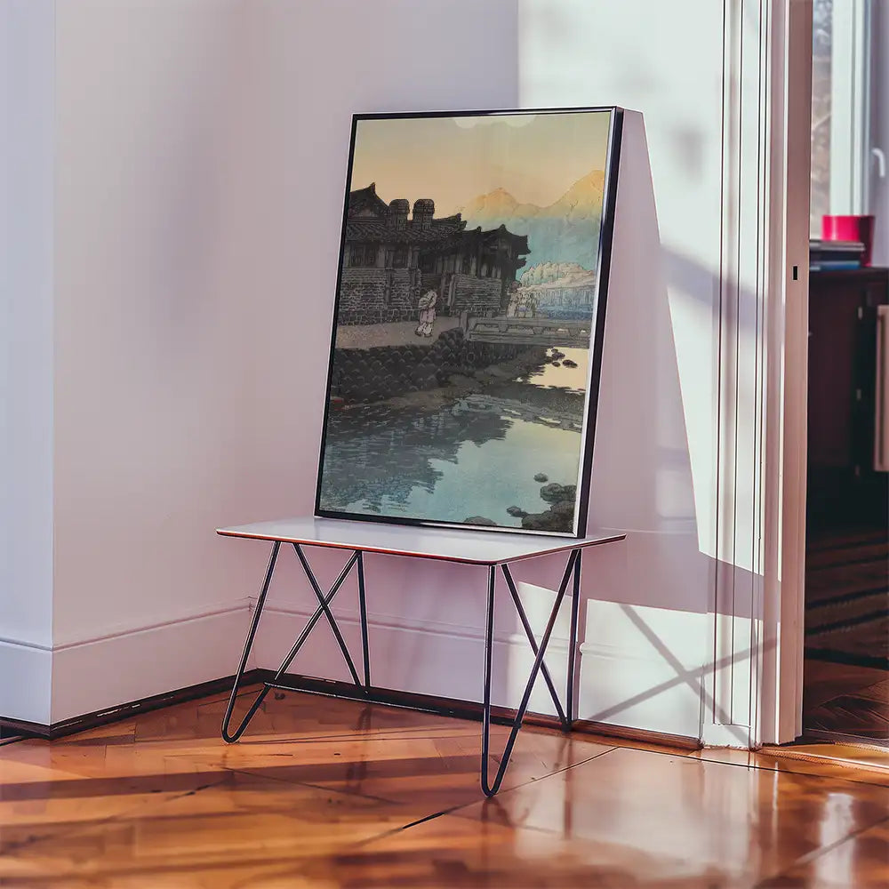 Framed painting leaning against a wall on a modern table, depicting a serene landscape with a traditional building and mountains reflected in water.